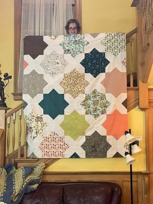 Woollypetals Floored quilt displayed on yellow wall with geometric star blocks