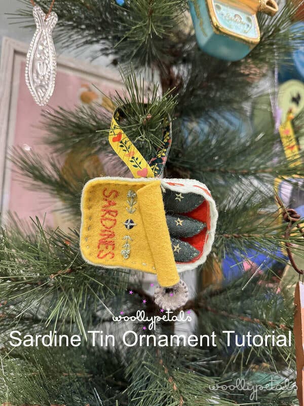 Woollypetals Sardine Tin Ornament in yellow felt with red text hanging on evergreen branch