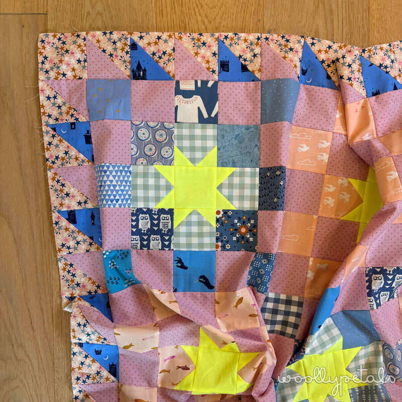 Woollypetals New Star quilt top with pink, blue, and neon yellow star blocks laid out on wooden floor